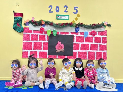 Seven cute Toddlers sitting in a row wearing pajamas in front of a paper fireplace art project.